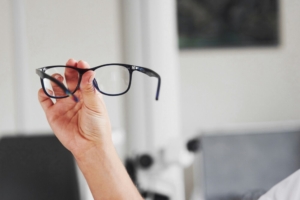 Hand holding black eyeglasses for inspection in a clean indoor setting, representing professional eyeglass frame assessment and repair.