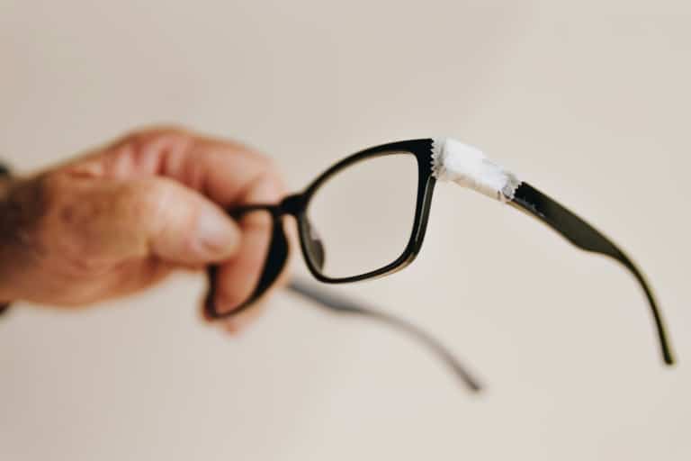 Hand holding black eyeglasses with white tape wrapped around the upper frame corner, showing a temporary fix for a broken glasses frame.