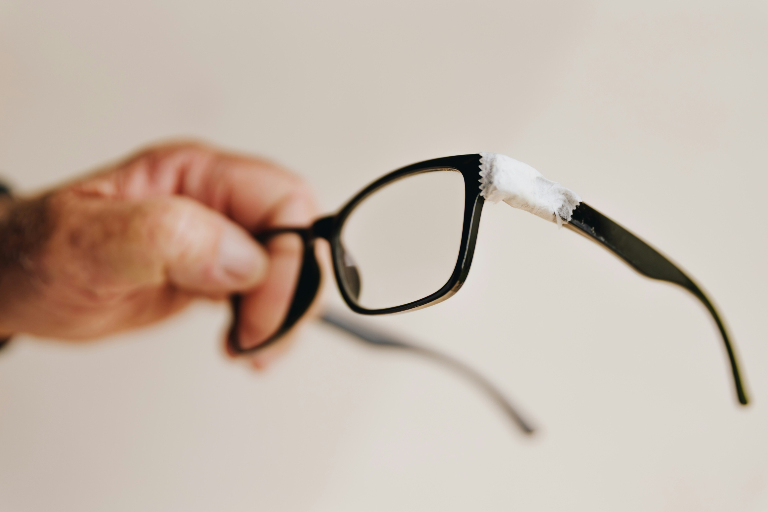 Hand holding black eyeglasses with white tape wrapped around the upper frame corner, showing a temporary fix for a broken glasses frame.