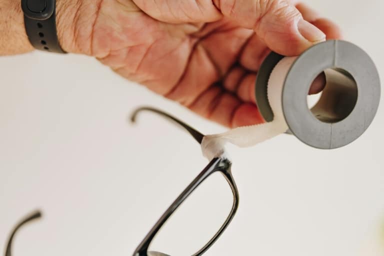 Close-up of a hand wrapping white tape around the temple area of black eyeglasses, showing a temporary fix for a broken glasses frame.
