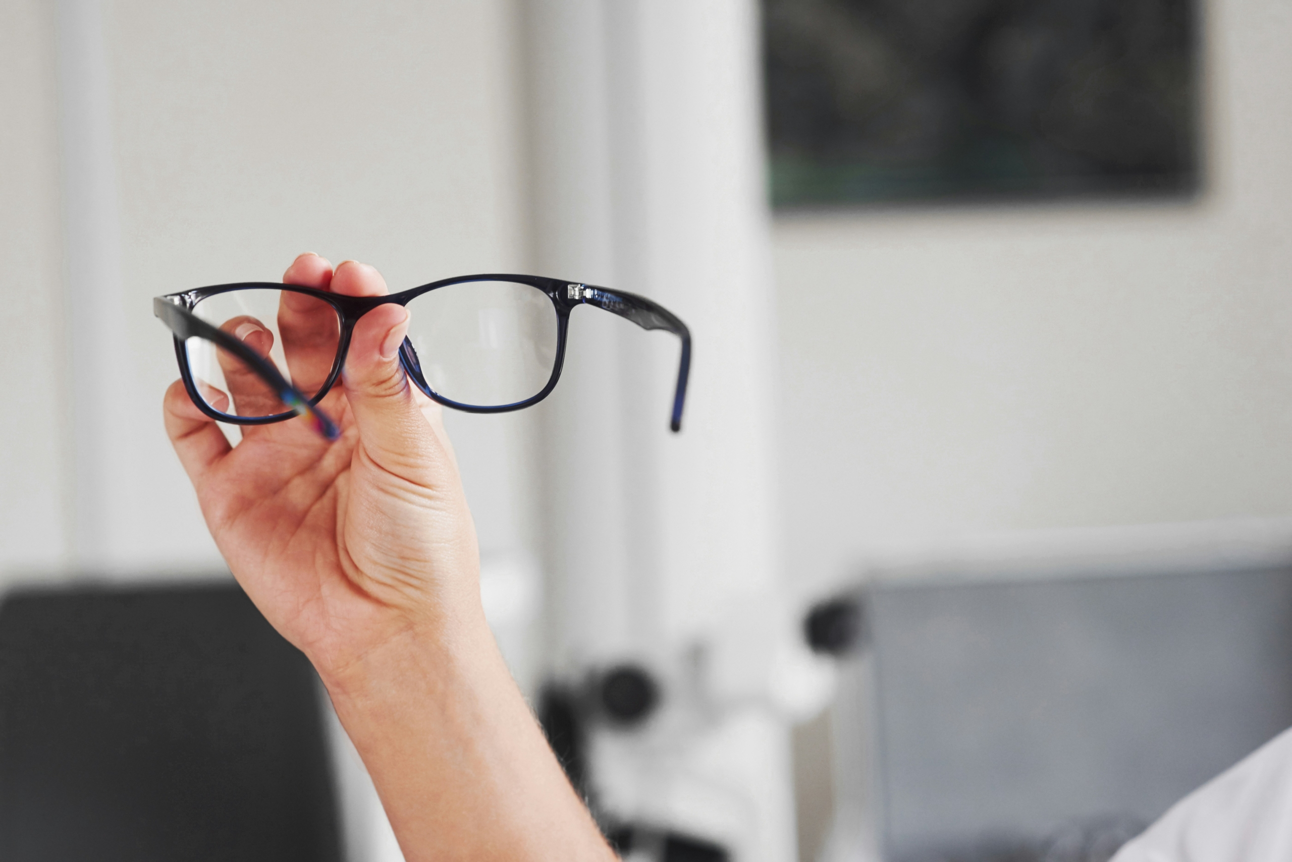 Hand holding black eyeglasses for inspection in a clean indoor setting, representing professional frame assessment and repair.