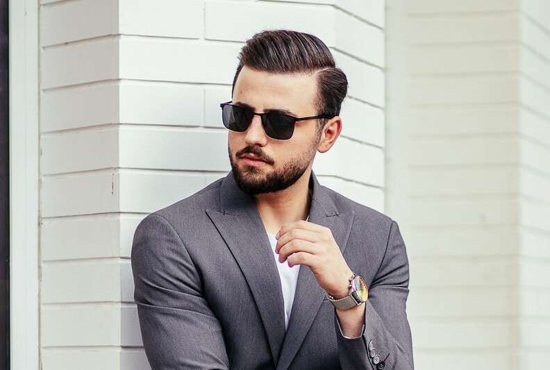 man in a suit wearing sunglasses A man wearing a grey blazer and dark sunglasses leaning against a white brick wall.