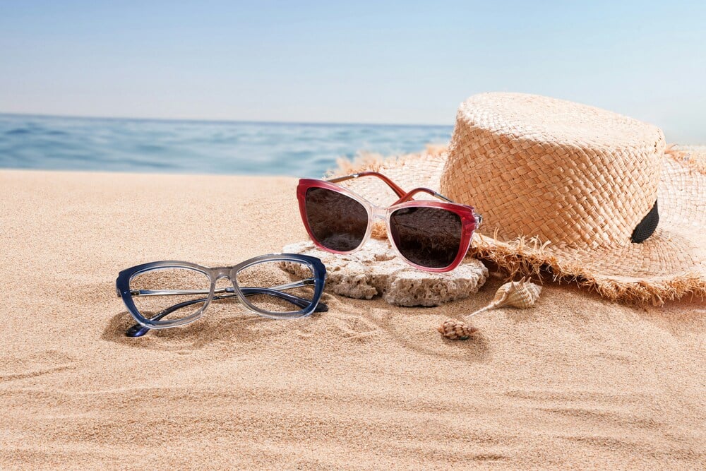 sunglasses by the beach Two pairs of sunglasses and a straw sun hat resting on a sandy beach next to the ocean.